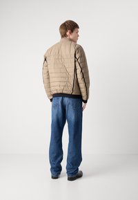 Casaco puffer bege acolchoado com colarinho alto, detalhes em preto e bainha canelada, combinado com jeans azuis de corte solto e sapatos pretos.