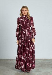 Largo vestido burdeos con estampados florales en rosa y blanco, corsé fruncido, mangas abullonadas y una falda fluida. Lleva sandalias negras.