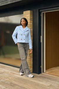 Hellblauer Wollmischung, doppelt geknöpfter Blazer mit goldenen Knöpfen, kombiniert mit grauen Schlaghosen und weißen Sneakers, auf einem Holzdeck.