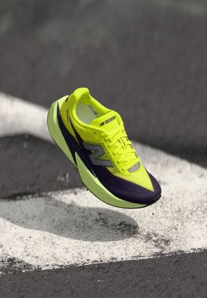 Scarpa da corsa giallo neon con accenti viola, tomaia leggera in mesh, design con lacci e suola lavorata, sospesa in aria sopra una linea dipinta.