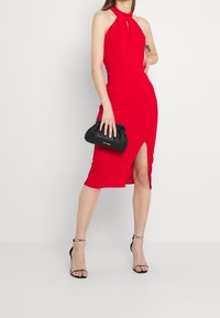 Röd midi klänning med halterneck och nyckelhålsdetalj fram, med en sidospalt. Kombineras med en svart kuvertväska och sandaler med remmar.