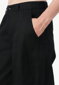 Main dans la poche d’un pantalon noir taille haute avec bouton et passants de ceinture, visible de la taille à la partie supérieure de la cuisse.