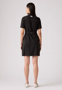 Zwart spijkerjurk met korte mouwen, een aangesloten taille, zichtbare stiksels en een riemdetail. De jurk heeft een kraag en een rechte zoom.