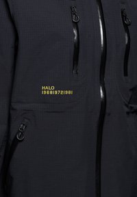 Sort jakke med tekstureret stof, tre lynlås-lommer og gul broderet tekst, der læser "HALO 196819721981."