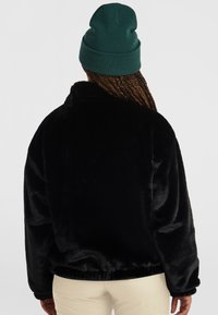 Zwarte nepbontjas met een hoge kraag, oversized pasvorm en elastische manchetten, gedragen met een groene gebreide beanie. Zachte textuur en warme uitstraling.