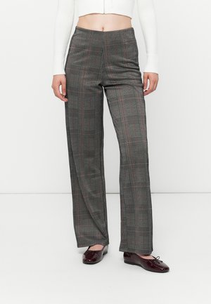 Vrouw draagt grijze geruite broek met hoge taille en wijde pijpen, gecombineerd met bordeauxrode platte schoenen en een kort wit shirt met lange mouwen.