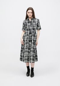 Rochie cămașă în carouri alb-negru, cu mâneci scurte, nasturi în față, talie strânsă și fustă fluidă. Stilizată cu cizme negre până la gleznă.