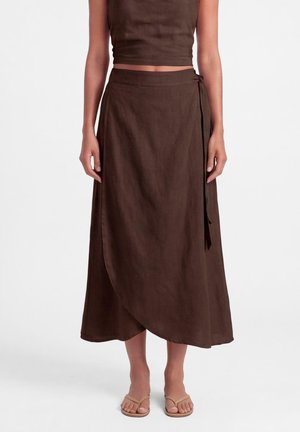 Vrouw draagt een bruine wikkel midi rok en bijpassende mouwloze crop top, staand op beige sandalen met blote voeten tegen een witte achtergrond.