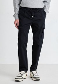 Black cargo pants with elastic waistband and drawstring. Features side pockets and a relaxed fit. Paired with white and green sneakers.