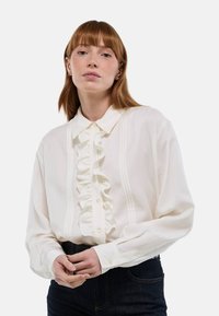 Blouse blanche à manches longues avec un devant à volants, fermeture à boutons et col structuré. Présente des détails de pinces discrètes le long de la patte de boutonnage.