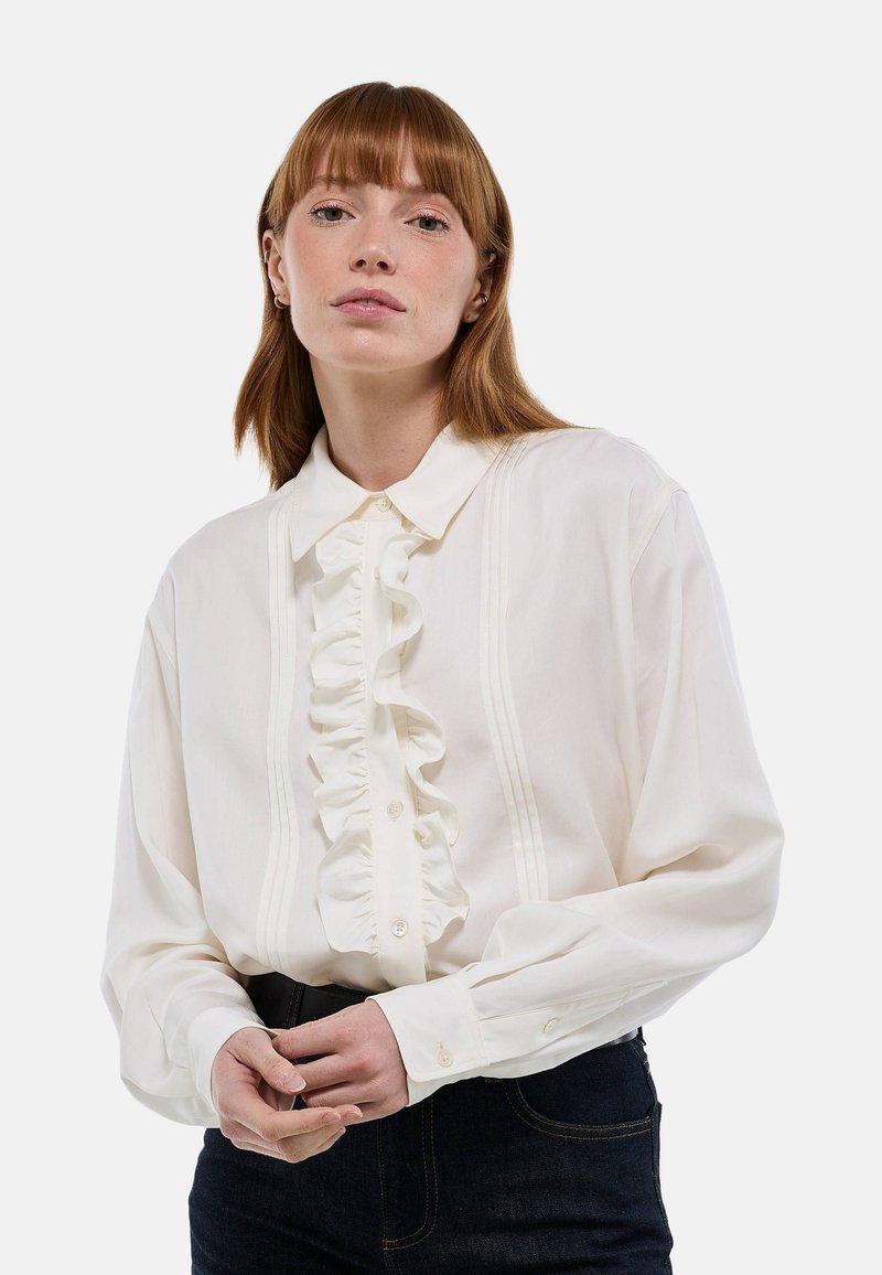 Blouse blanche à manches longues avec un devant à volants, fermeture à boutons et col structuré. Présente des détails de pinces discrètes le long de la patte de boutonnage.