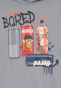 Impression graphique sur un hoodie gris mettant en avant quatre cans de spray de différentes couleurs avec des designs de singes de cartoon et le texte "RESTEZ ENNUYÉ."