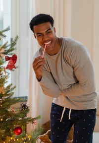 Grijs shirt met lange mouwen, marineblauwe broek met candycane-print, glimlachend terwijl hij een rood-wit gestreepte candycane vasthoudt naast een versierde kerstboom.