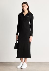 Μαύρο μακρυμάνικο πόλο φόρεμα με γιακά και πράσινο λογότυπο. Συνδυασμένο με λευκά αθλητικά παπούτσια και μια μικρή μαύρη τσάντα.