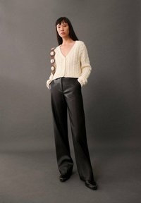 Elfenben farvet kabelstrik cardigan med V-hals og fem runde knapper på et brunt bånd, parret med højhalset sorte læderbukser.