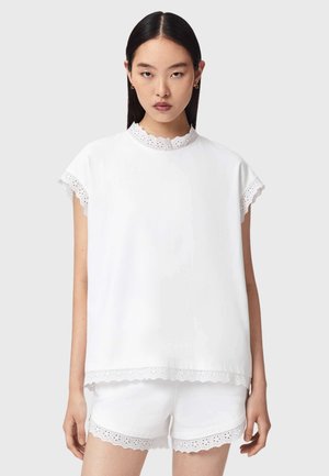 Femme aux cheveux raides et foncés portant un ensemble short et haut à manches courtes blancs avec une bordure en dentelle ajourée à bords festonnés.
