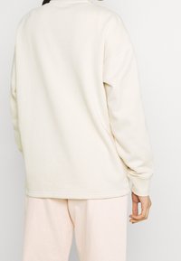 Krämfärgad sweatshirt med avslappnad passform, rund halsringning och ribbade ärmslut, tillsammans med ljusrosa byxor. Mjuk tygtextur.