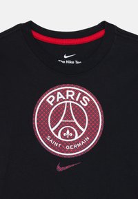 Melna Nike T-krekls ar sarkanu un baltu Paris Saint-Germain logotipu un mazu saderīgu Nike swoosh apakšā.