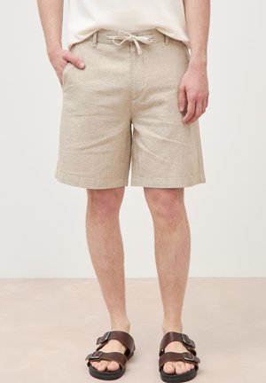 Mann trägt beige Shorts mit Kordelzug, Hand in der Tasche und dunkelbraune Sandalen mit Doppelriemen, steht auf hellem Boden.
