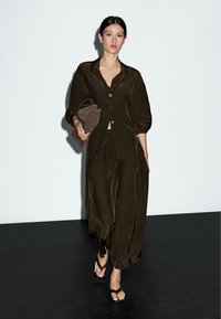 Woman walking in loose dark brown satin outfit with wide-leg pants and buttoned shirt, carrying a matching clutch and wearing dark sandals.