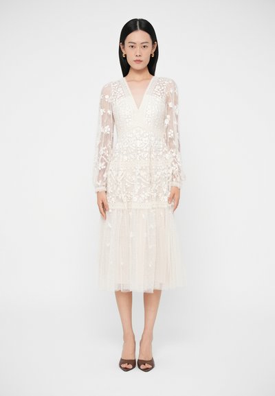 Donna che indossa un abito midi bianco in pizzo floreale con maniche lunghe trasparenti e scollo a V, abbinato a décolleté scuri con punta affusolata, in piedi davanti a uno sfondo semplice.