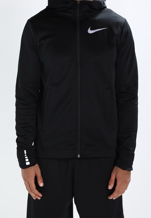 Personne portant un sweat à capuche zippé noir Nike avec une capuche et un logo réfléchissant sur la poitrine et le poignet, debout, les mains détendues le long du corps.