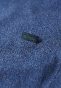 Blauwe stof met een zachte textuur; heeft een klein zwart label met groene tekst "Superdry." Eenvoudig en uniform patroon.