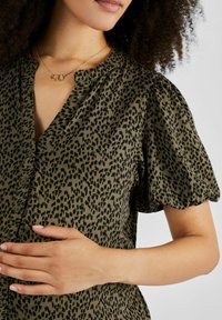 Zelenošedá košeľa s čiernym leopardím vzorom, s krátkymi puff rukávmi a gombíkovým zapínaním. Textúrovaná tkanina s jemným leskom. Zlatý náhrdelník s prepletenými článkami.