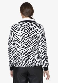 Donna con coda di cavallo marrone che indossa un maglione a strisce zebra bianco e nero e pantaloni neri, voltata di spalle contro uno sfondo bianco semplice.