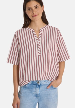 Femme portant un chemisier blanc à manches courtes avec des rayures rouges verticales et un jean bleu clair, debout avec une main dans la poche devant un fond uni.