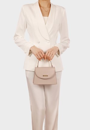 Donna con blazer doppiopetto bianco e pantaloni che tiene una piccola borsa beige con manico superiore e targa con logo dorato.