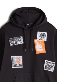 Sudadera negra con diversos parches en naranja y blanco. Los parches representan gráficos, incluyendo una cámara y diseños geométricos.