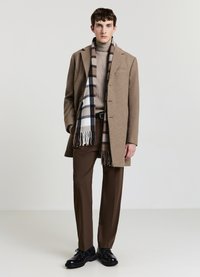 Brauner Wollmantel, gerippter beiger Rollkragenpullover, kariertes Halstuch in Beige und Schwarz, braune Hose, glänzende schwarze Schuhe. Minimalistisches Design, maßgeschneiderte Passform.