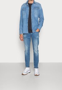 Ljusblå denimskjorta med två bröstfickor, bärs över en mörk t-shirt; matchad med blekta blå jeans och vita sneakers med bruna accenter.