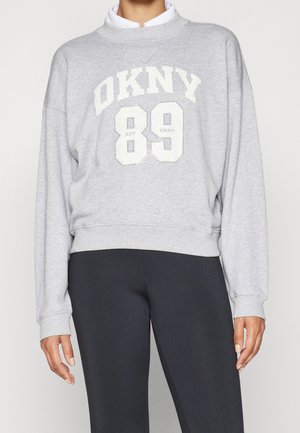 Person trägt einen grauen DKNY-Sweatshirt mit weißen Lettern und der Nummer 89, kombiniert mit schwarzen Hosen vor einem schlichten Hintergrund.