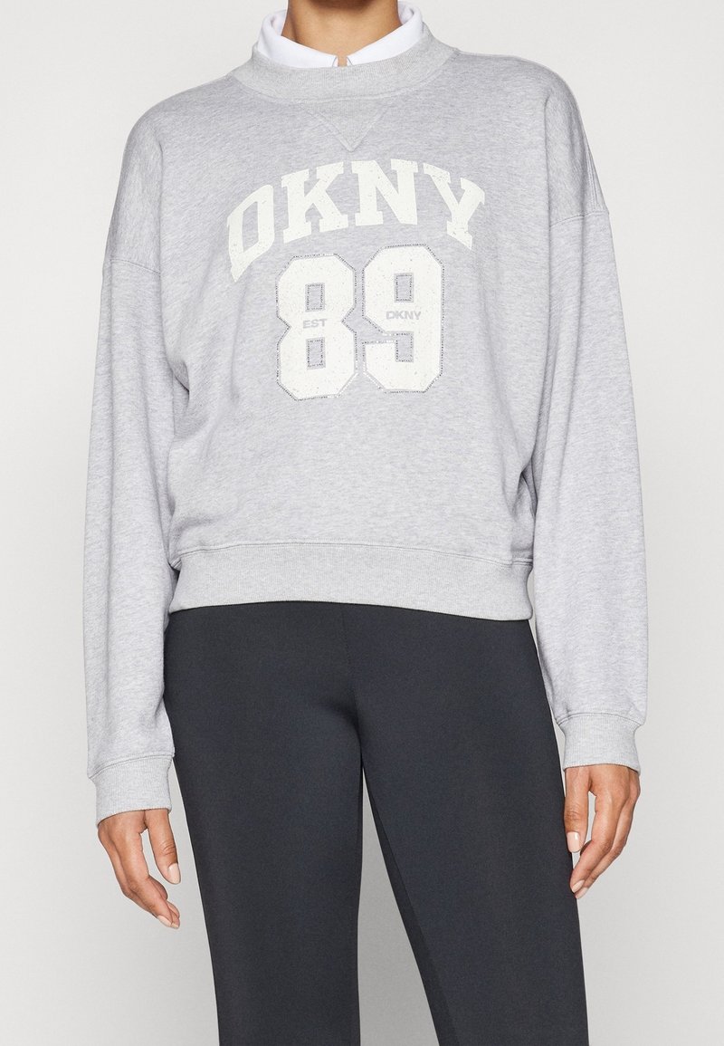 Personne portant un sweat-shirt gris DKNY avec lettres blanches et numéro 89, associé à un pantalon noir sur un fond uni.