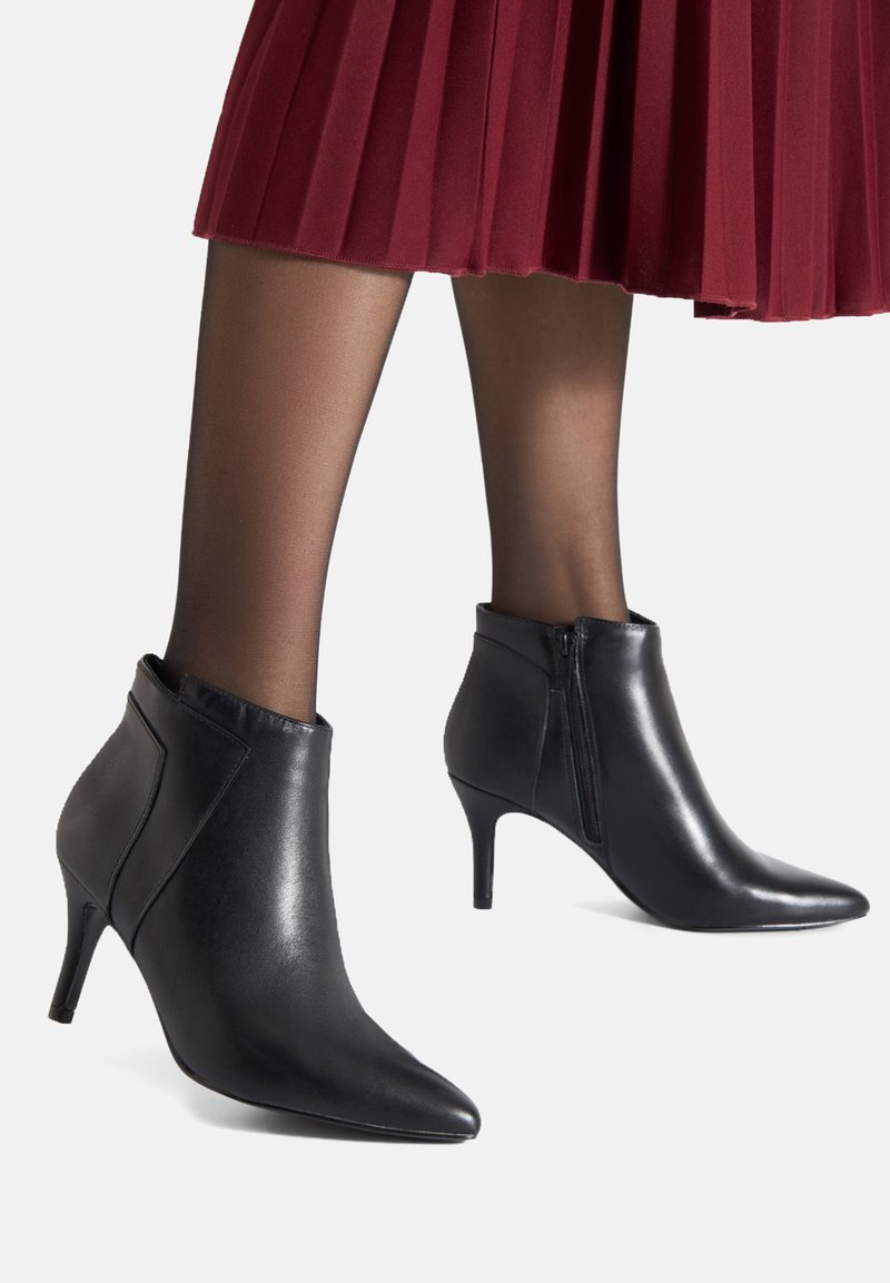 Botines de cuero negro con puntera puntiaguda y tacón de 3 pulgadas; cuentan con una cremallera lateral y una textura suave. Combinados con una falda plisada.