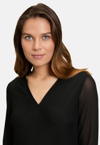Zwarte V-hals top met lange doorzichtige mouwen, gladde textuur, zachte stof die valt en een subtiele glans tegen een effen witte achtergrond.