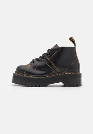 Bottine plateforme en cuir noir avec surpiqûres jaunes, semelle épaisse à rainures, laçage devant et languette avec logo au talon.