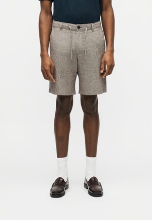 Homme portant un short beige à cordon, des chaussettes blanches montantes, des mocassins marron foncé et une chemise bleu marine foncé, debout devant un fond blanc uni.