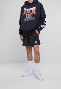Sudadera negra con el logo "STARTER" en rojo y blanco, pantalones cortos de malla grises y zapatillas blancas. Presenta puños acanalados y acentos deportivos.