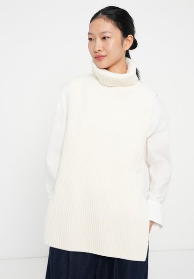 Rebrasti, krem boje turtleneck prsluk od debelog pletenog materijala, uparen s bijelom košuljom na gumb i tamnoplavom suknjom.