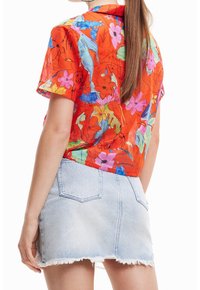 Femme portant une chemise rouge à fleurs à manches courtes et une mini-jupe en jean bleu clair effilochée, vue de dos avec de longs cheveux bruns.
