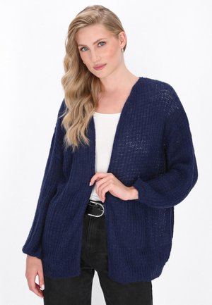 Cardigan de culoare bleu marin, din tricot deschis, realizat dintr-un material texturat. Dispune de umeri lăsați și mâneci lungi, purtat deasupra unei bluze albe și a unor pantaloni negri.