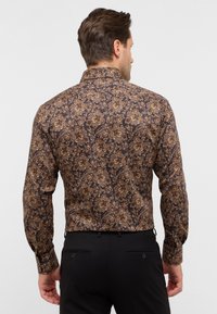 Camicia a maniche lunghe marrone con motivo floreale, dotata di colletto e polsini con bottoni, abbinata a pantaloni neri.