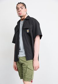 Cămașă neagră cu mânecă scurtă, cu un buzunar pe piept, purtată peste un tricou gri deschis, asortată cu pantaloni scurți kaki, din pânză, cu buzunare laterale.