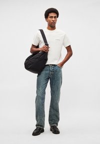 Biele bavlnené tričko s malým čiernym textom, modré džínsové nohavice, čierna kabelka na rameno a čierne robustné topánky, nosené stojacim mužským modelom.