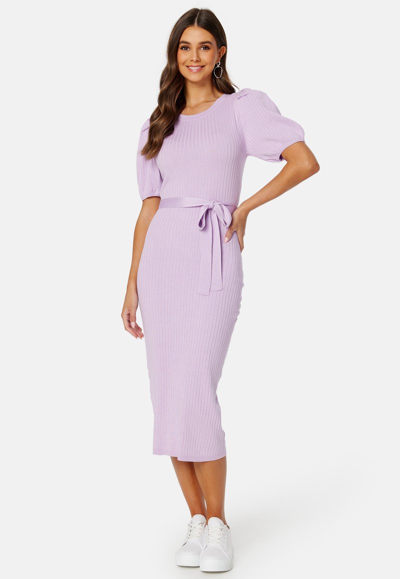 Abito a coste, lungo fino al ginocchio, in lavanda con maniche a palloncino corte e cintura annodata in vita. Abbinato a sneaker bianche.