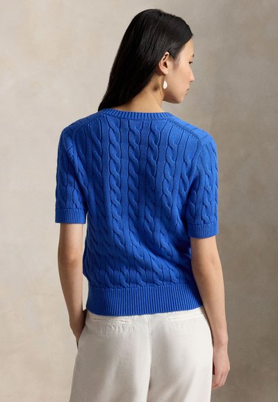 Ženska, oblečena v modri kratek pulover s kablasto pletenino in bele hlače, prikazana od zadaj na nevtralnem ozadju.