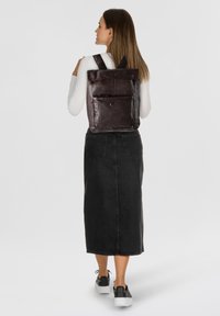 Zaino in pelle di colore marrone scuro con design a risvolto, tasca frontale, spallacci regolabili e texture liscia. Indossato da un modello.